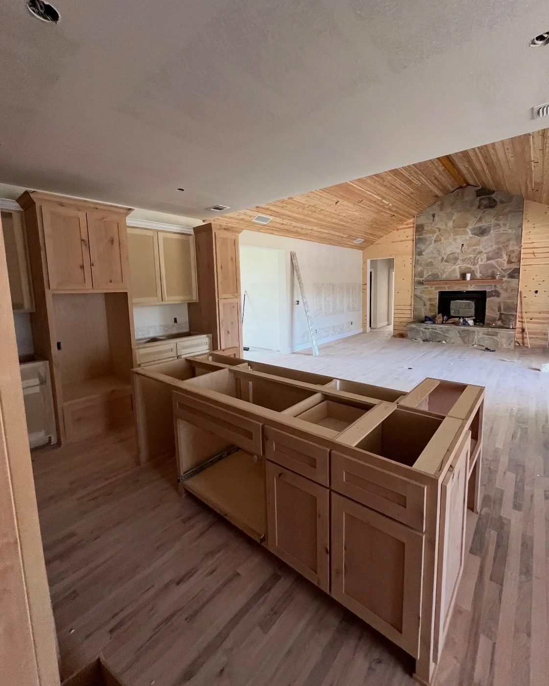 Unfinished kitchen island and surrounding wood cabinetry in an open room with a stone fireplace.